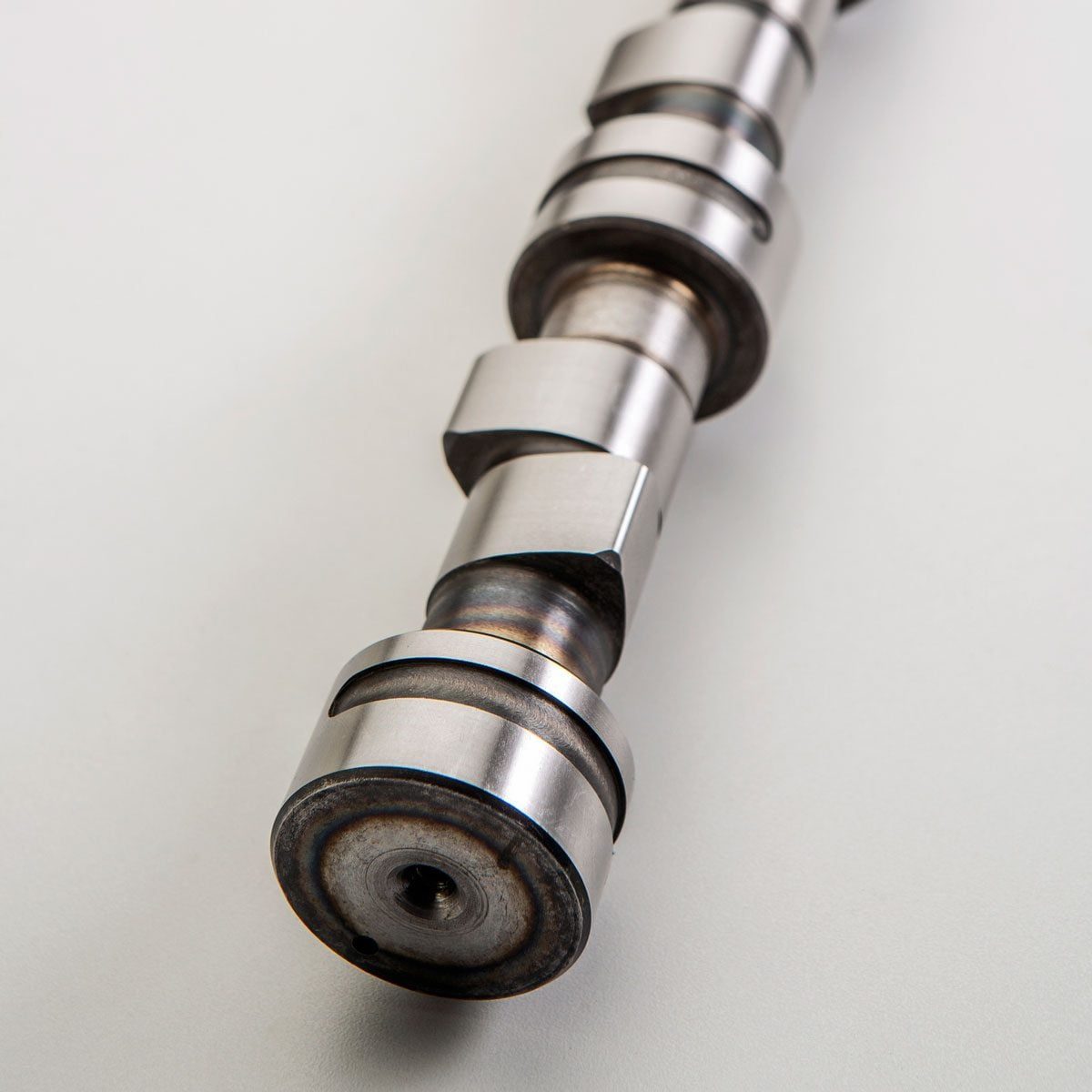 A metallic camshaft lies on a light surface, showcasing its cylindrical form and multiple grooves, suggesting functionality in an engine sys