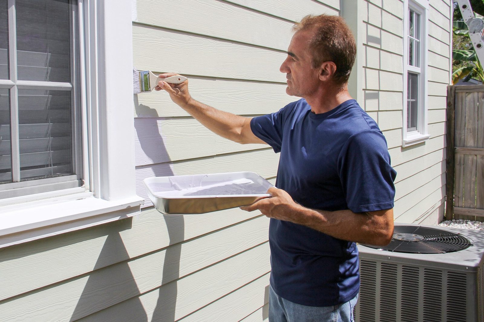A man paints the trim of a house with a brush while holding a paint tray, positioned next to an air conditioning unit in a sunny environment