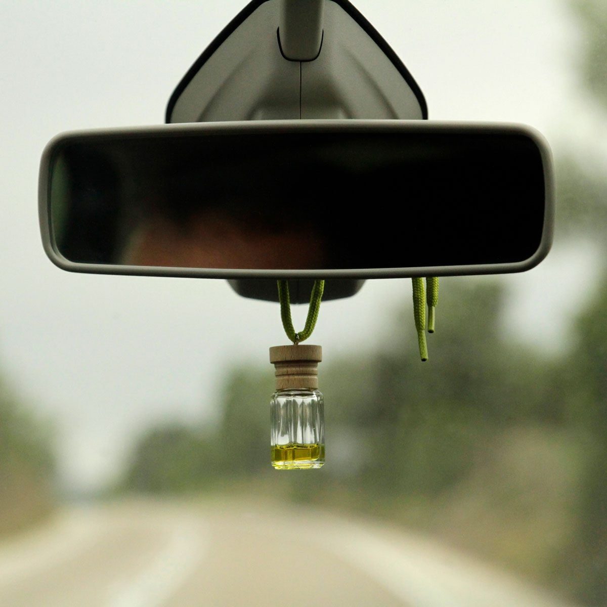 A glass bottle filled with yellow liquid hangs from a mirror in a car, with a blurred road and greenery visible in the background.