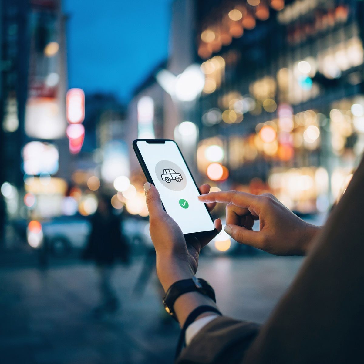 A hand holds a smartphone, displaying a car icon and checkmark, while in a brightly lit urban environment during twilight.