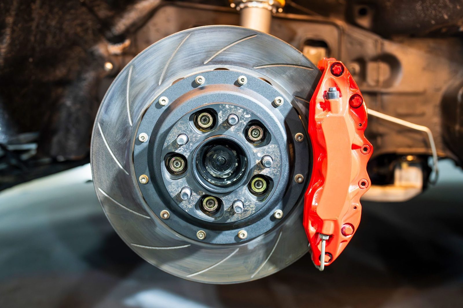 A red brake caliper grips a metallic brake disc, showcasing detailed components in a well-lit automotive environment beneath a vehicle.