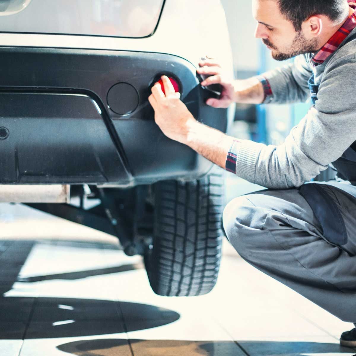 A mechanic inspects the rear of a car, focusing on a red light, in a well-lit garage with visible vehicle components and tiles.