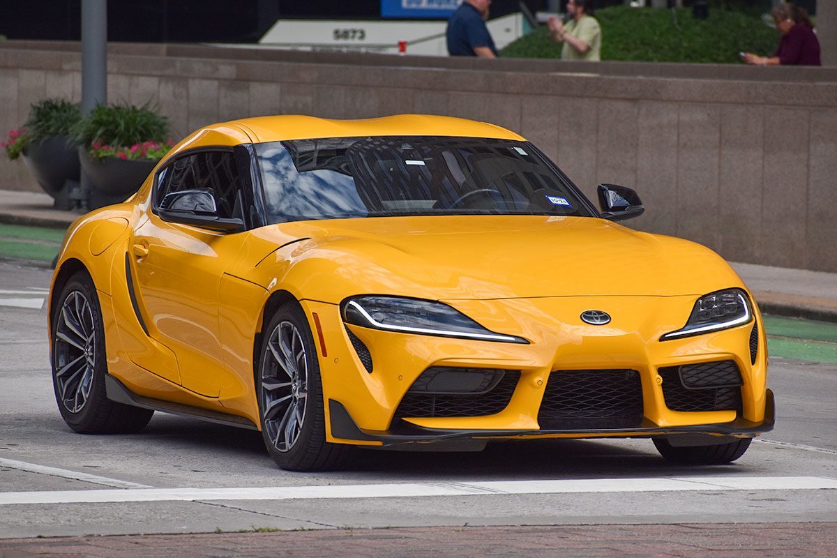 A portrait of a yellow Toyota GR Supra 2 sports car cruising in a downtown district