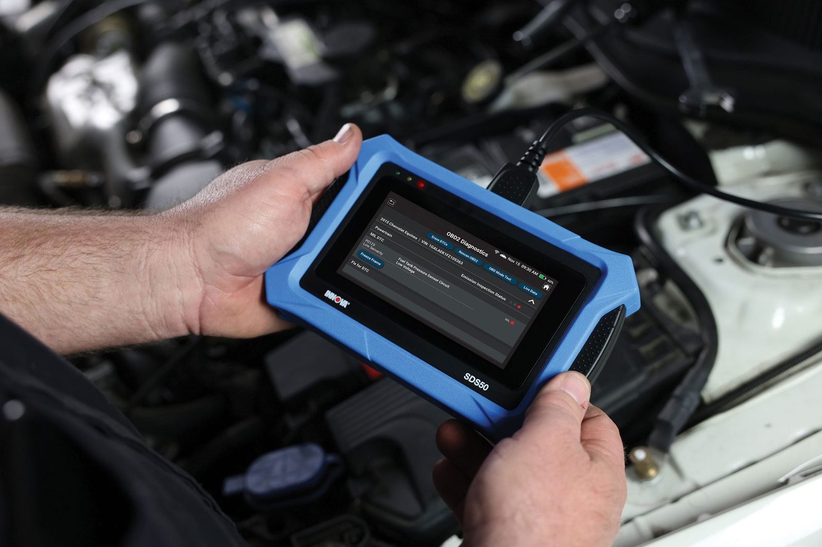 A handheld diagnostic tool displays information while a person holds it near a vehicle's engine bay, assessing diagnostic data and potential