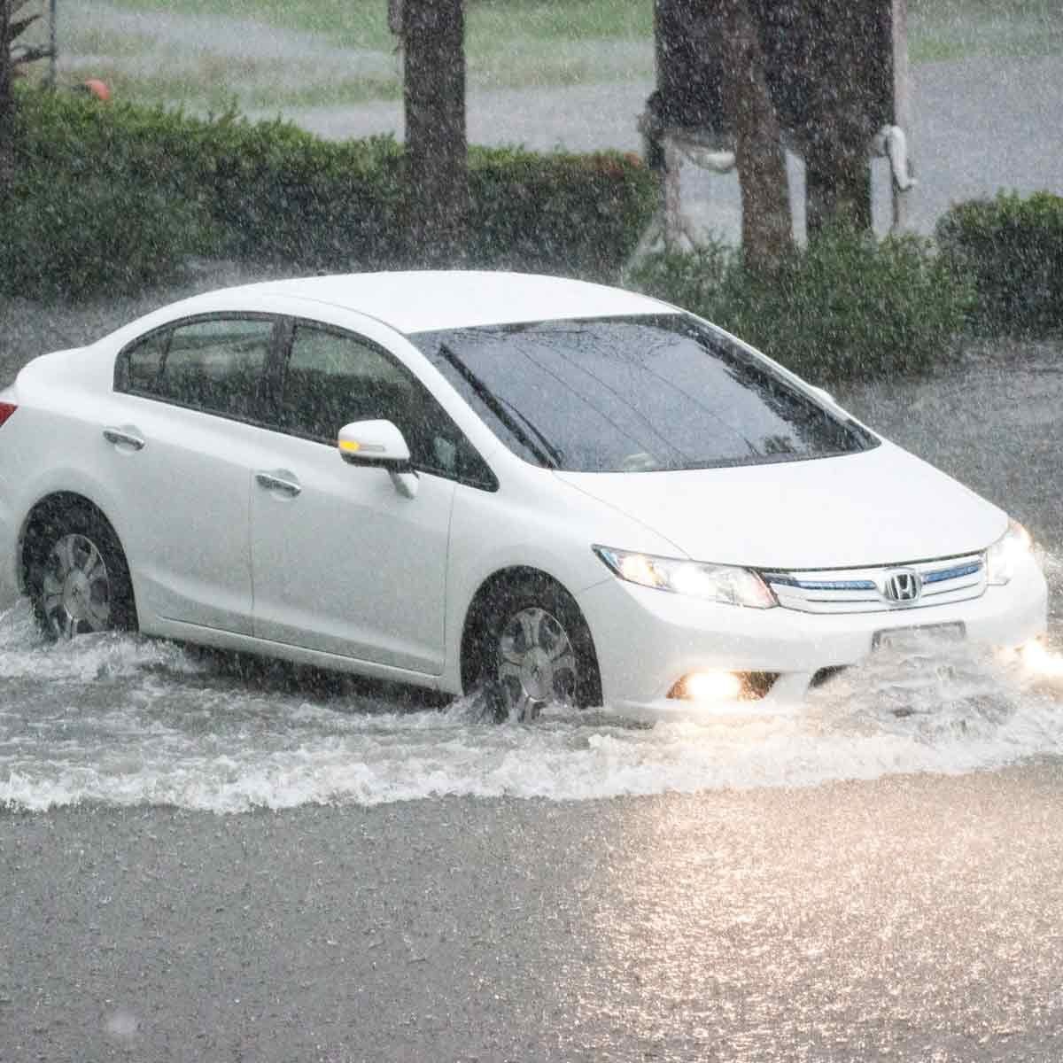 A white car drives through flooded streets, creating splashes as heavy rain falls, surrounding vegetation partially visible in the backgroun