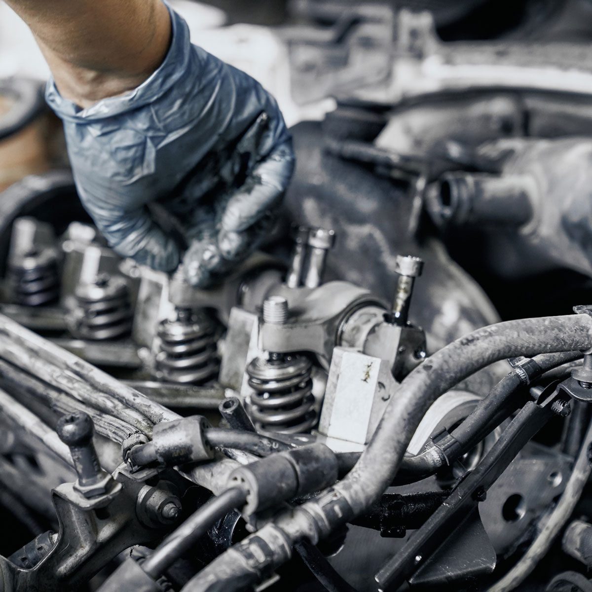 A gloved hand adjusts components in a greasy car engine, surrounded by mechanical parts and tools in a workshop environment.