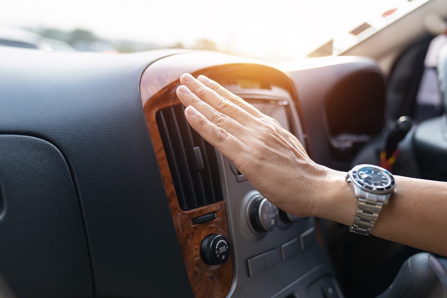 A hand adjusts the air vent controls in a car, set against a backdrop of a sunlit interior, indicating operation or comfort adjustments.