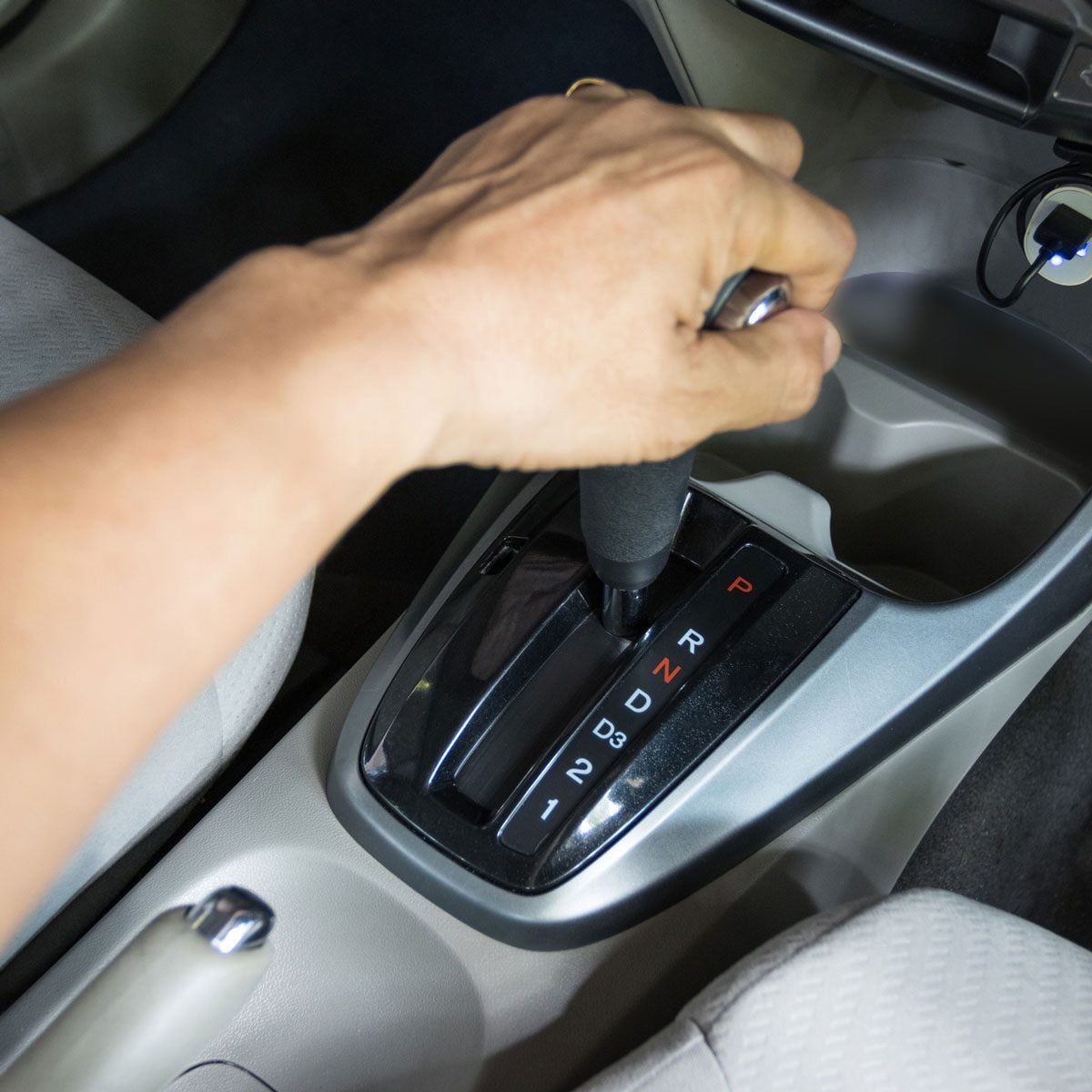 A hand adjusts the gear lever in a car, transitioning toward neutral, within a light-colored interior featuring textured seats and a simple