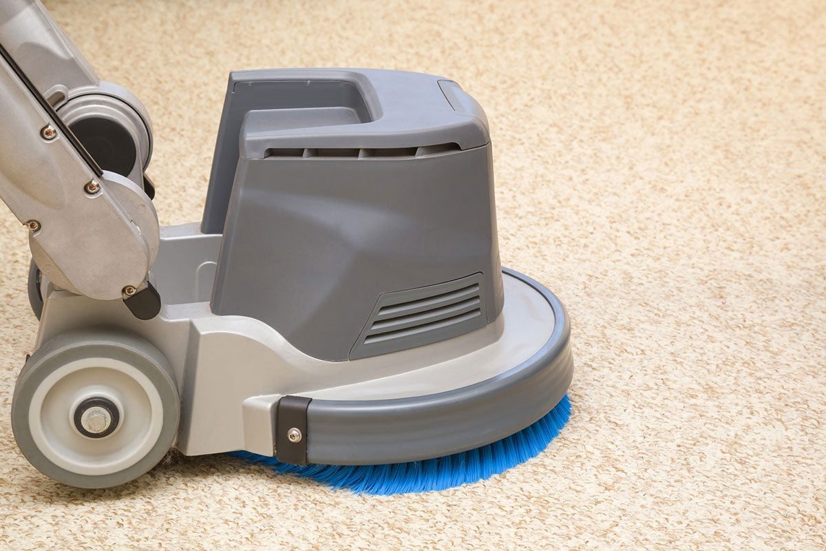 A floor cleaning machine operates, thoroughly scrubbing a light-colored carpet surface, surrounded by a clean indoor environment.