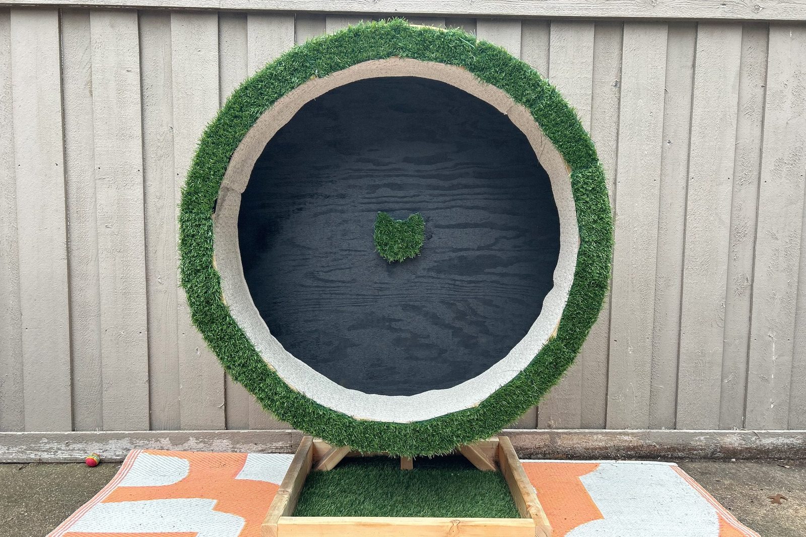 A circular structure with faux grass surrounds a dark, smooth interior. It's positioned near a wooden wall, on a patterned mat.