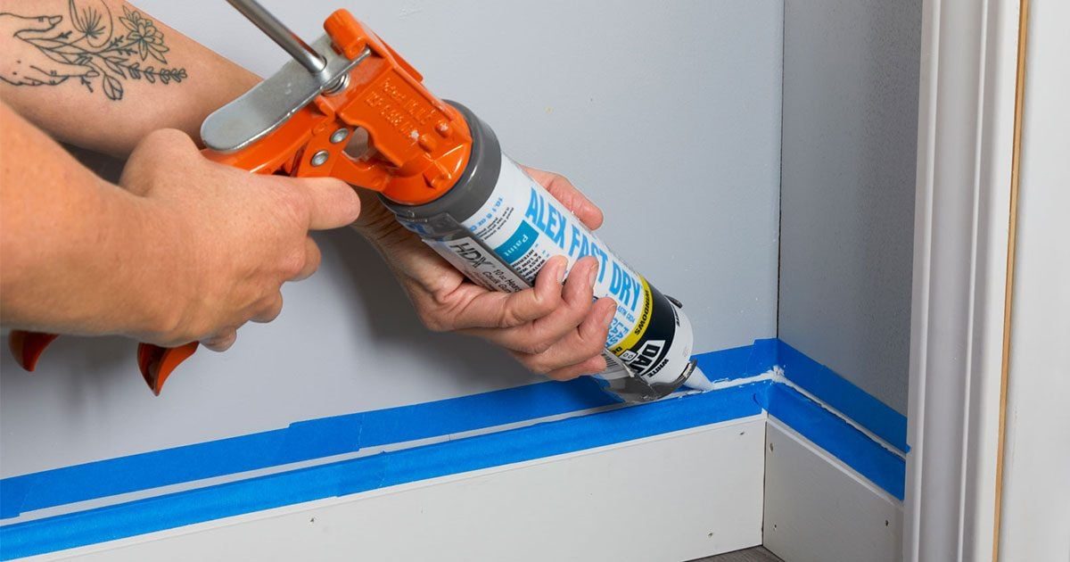 A hand applies caulk from a cartridge to a corner where blue painter's tape is positioned along a baseboard in a room.