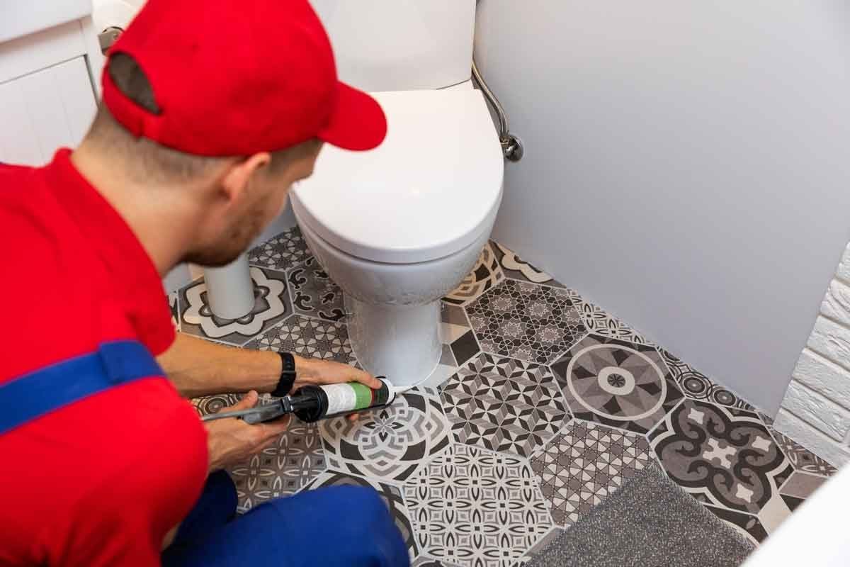 A worker applies sealant around a toilet's base, situated on a patterned tile floor in a bathroom. The worker wears a red shirt and cap.