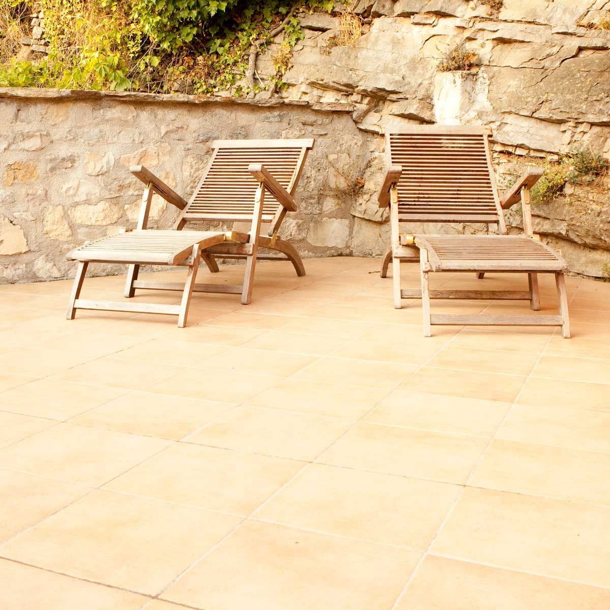 Two wooden lounge chairs are positioned on a tiled surface, set against a natural stone wall adorned with some greenery.