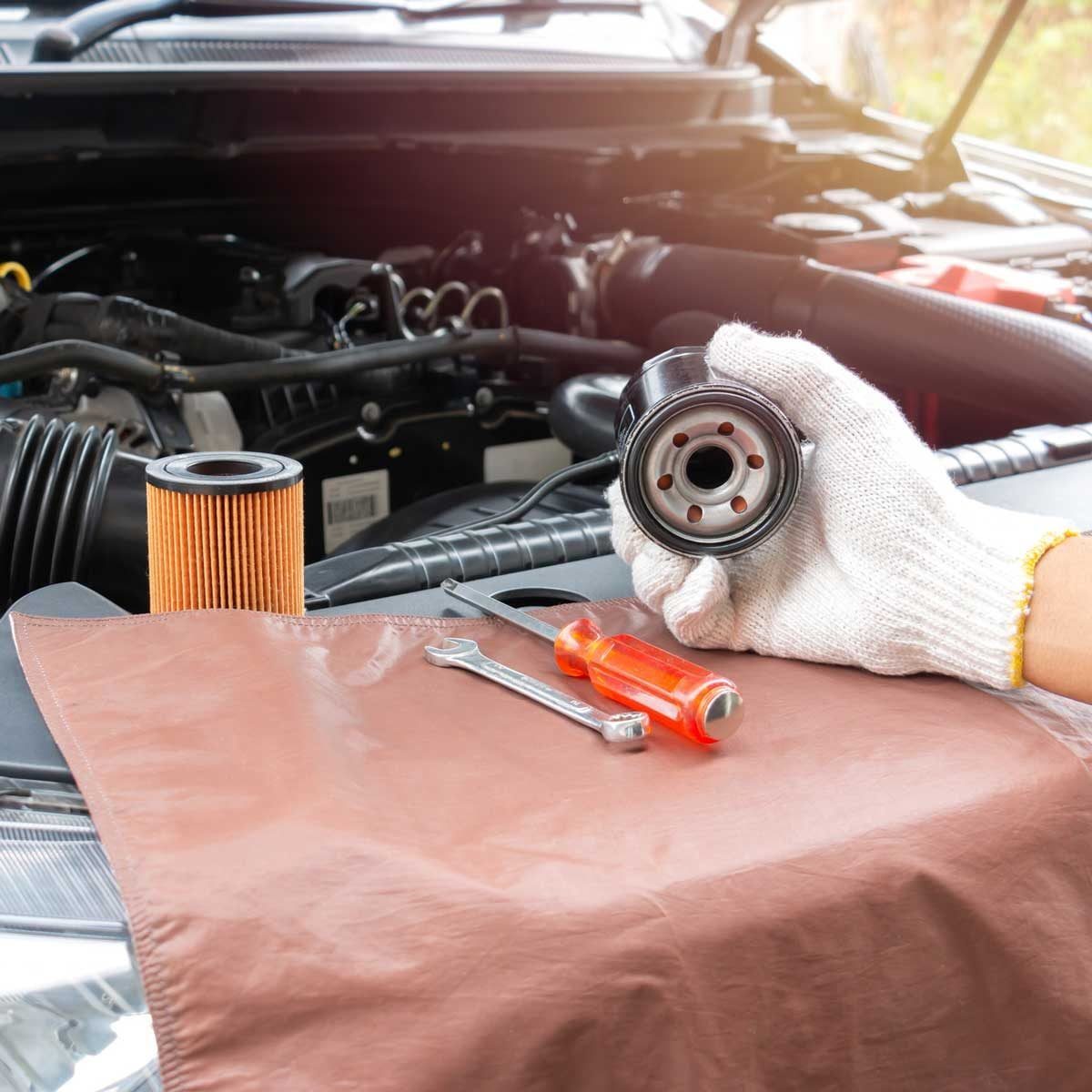 A hand in a glove holds an oil filter above a work surface with an orange oil filter, wrench, and screwdriver, while a car engine is visible