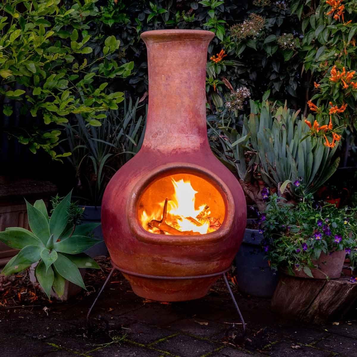 A clay chiminea emits flames, providing warmth. It's surrounded by green plants and colorful flowers, set against a dark, paved surface.