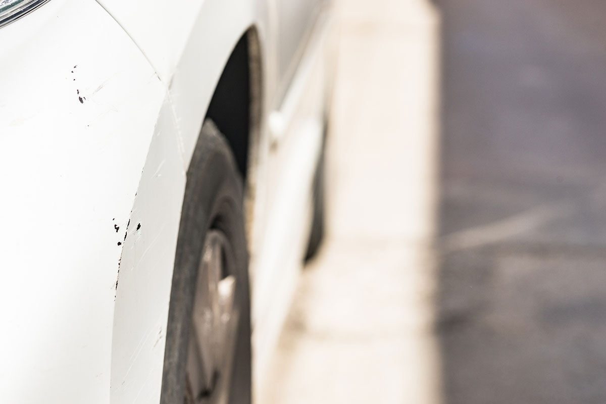 A white car’s wheel and fender are depicted, showing scratches and scuff marks, set against a blurred asphalt surface.