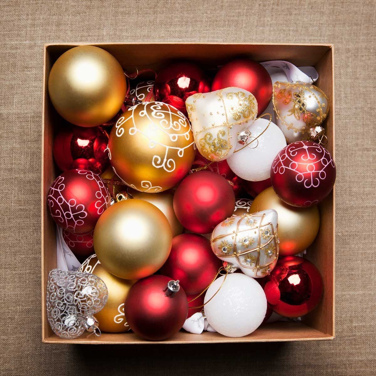 A box overflows with colorful Christmas ornaments, including red, gold, and white spheres, reflecting a festive atmosphere on a textured bac
