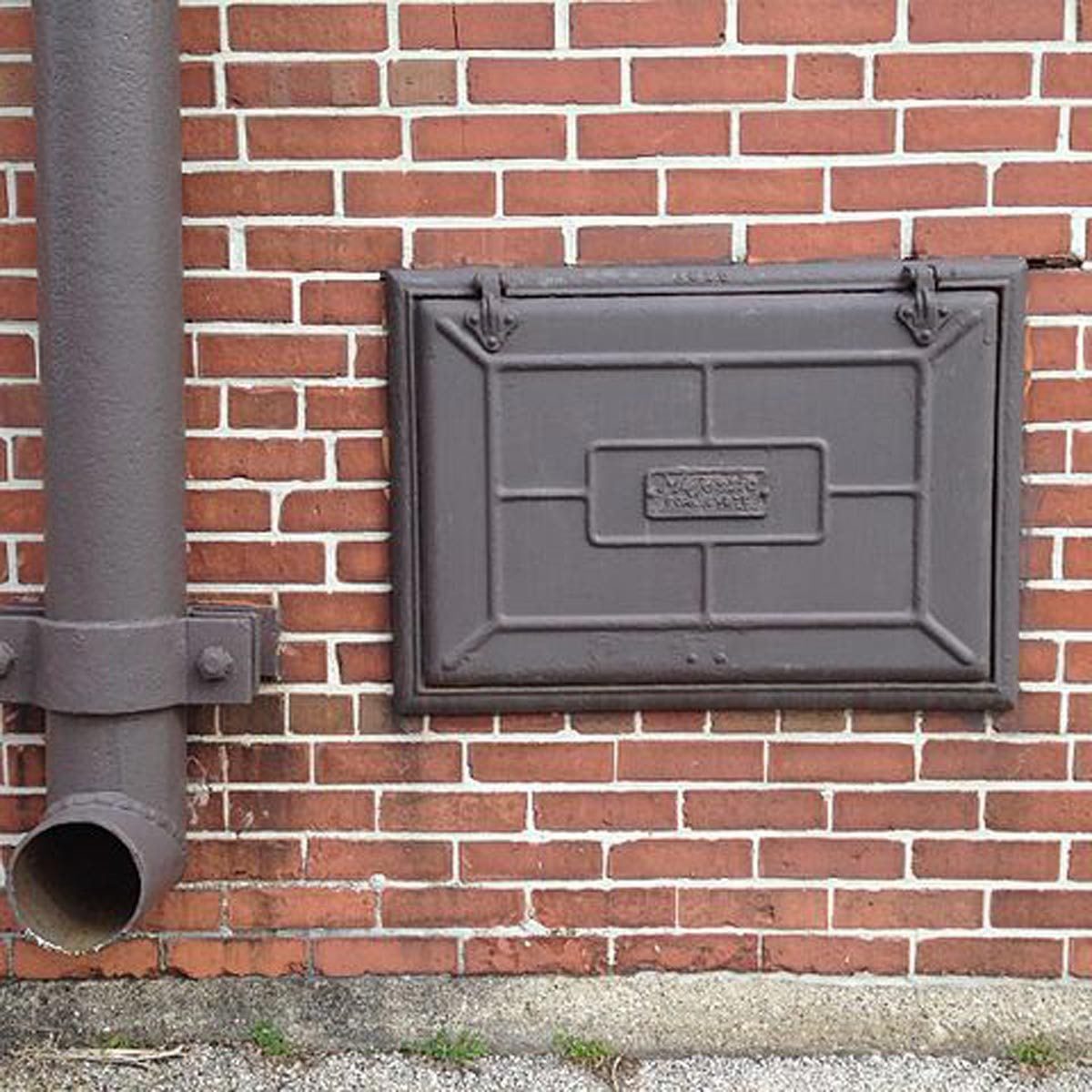 A rectangular, dark metal hatch is mounted on a red brick wall, next to a vertical pipe that has a circular opening at its bottom.