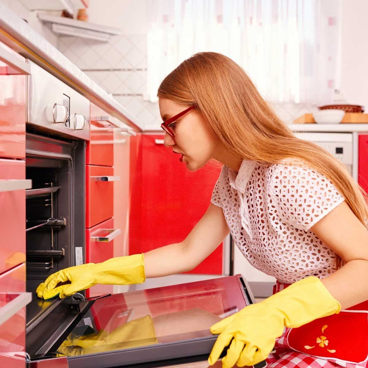 A woman wearing yellow rubber gloves cleans an oven in a vibrant red kitchen, focusing intently on the oven's interior.