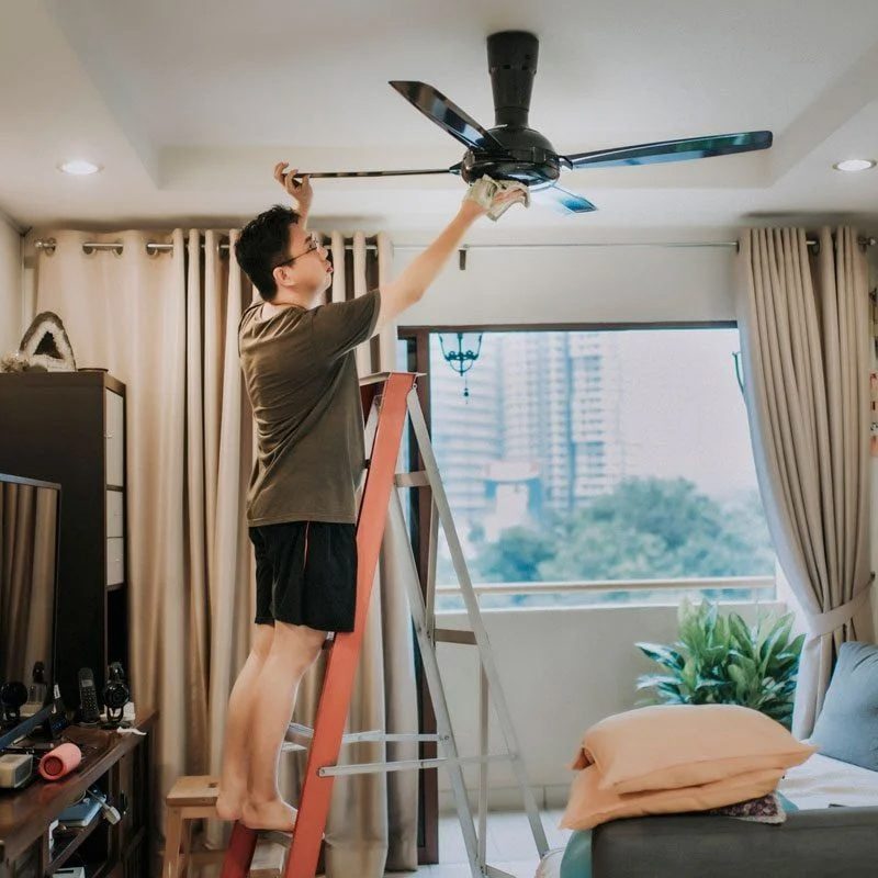 A person stands on a ladder, cleaning a ceiling fan in a living room filled with light, curtains, and a view of buildings outside.
