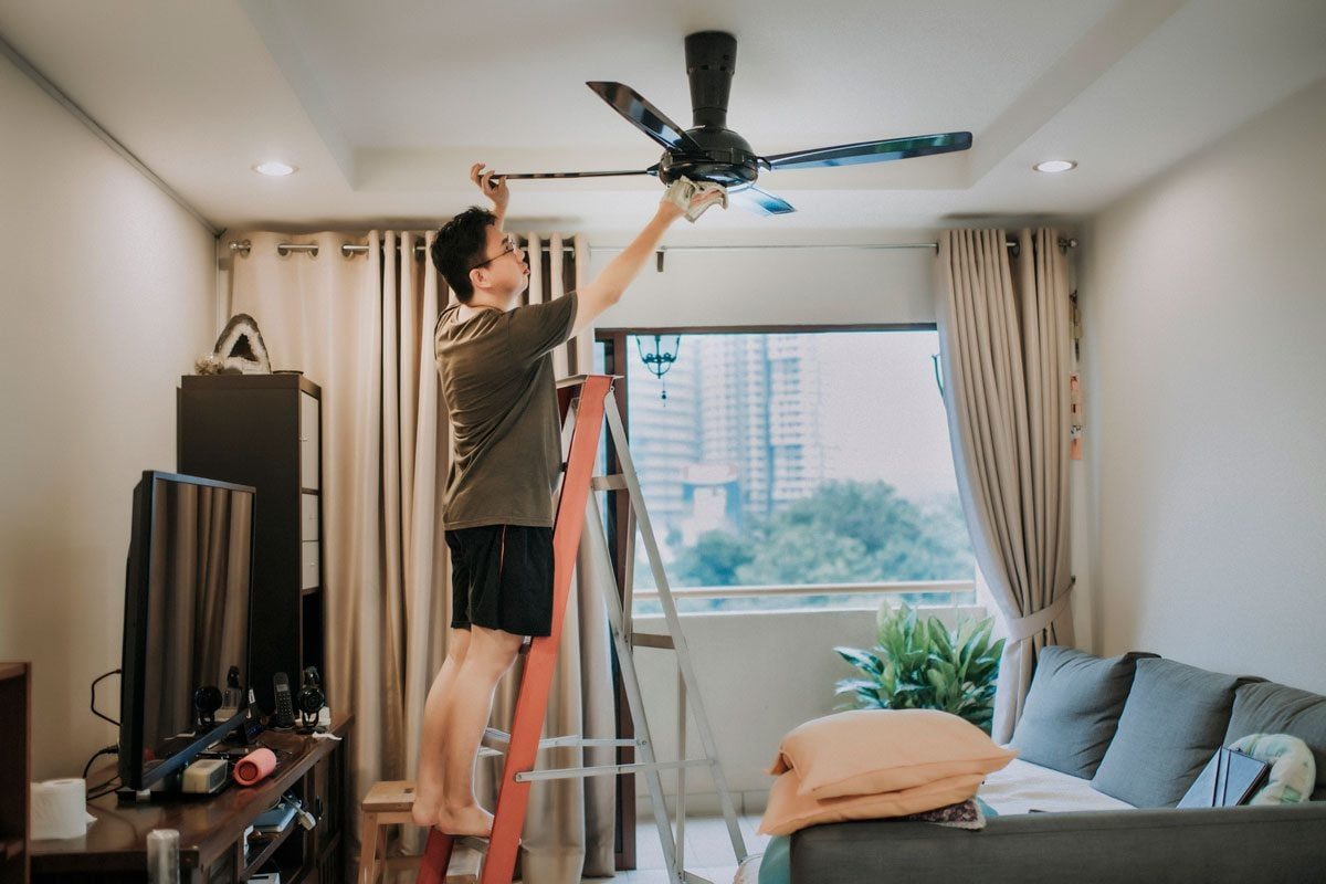 A person stands on a ladder, adjusting a ceiling fan in a living room with curtains, a couch, and a view of a cityscape outside.