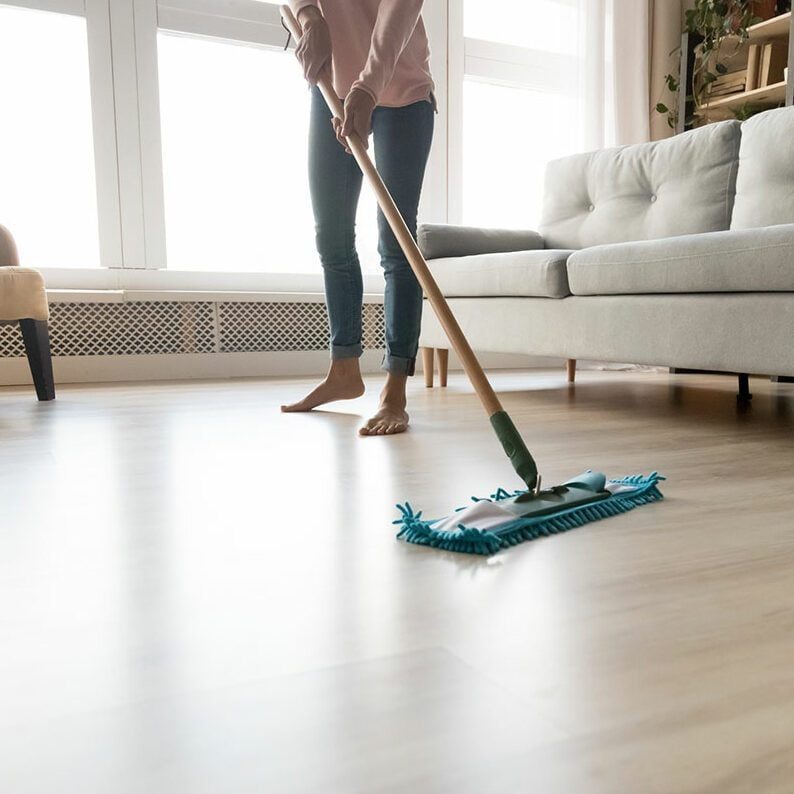 A person mops a wooden floor in a bright room with a gray couch and windows, enhancing the space's cleanliness and orderliness.