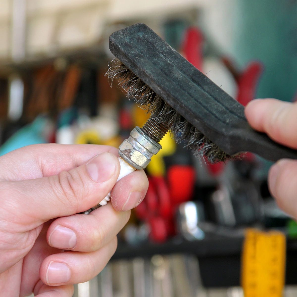 A hand holds a wire brush, cleaning threads of a metal object, surrounded by various tools in a workspace.