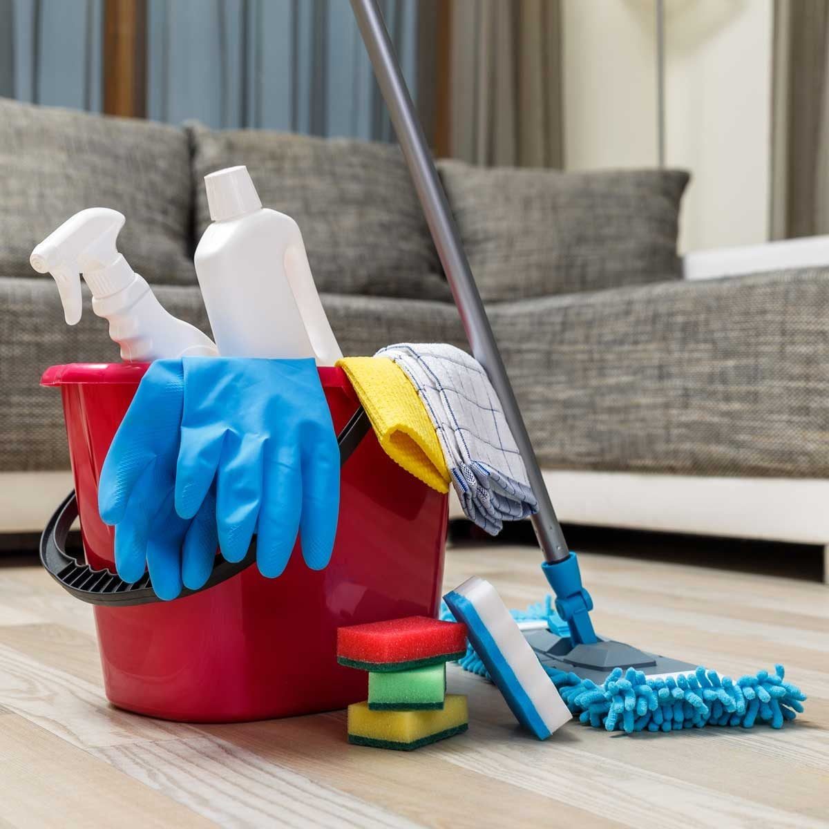 A red cleaning bucket holds sponges, a mop, and spray bottles, with blue gloves resting over the edge, positioned on a wooden floor near a c