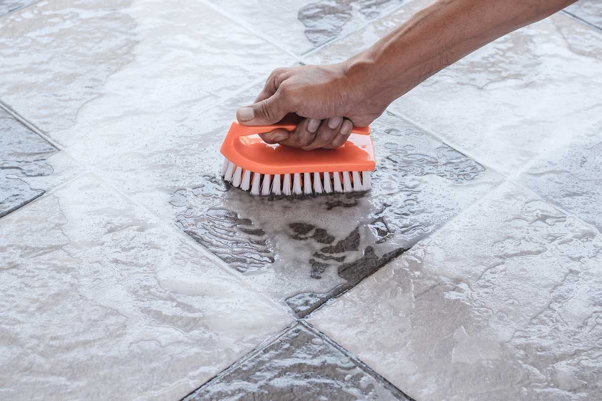 A hand holds a orange cleaning brush, scrubbing wet, tiled flooring marked with soap suds, in a domestic or maintenance setting.