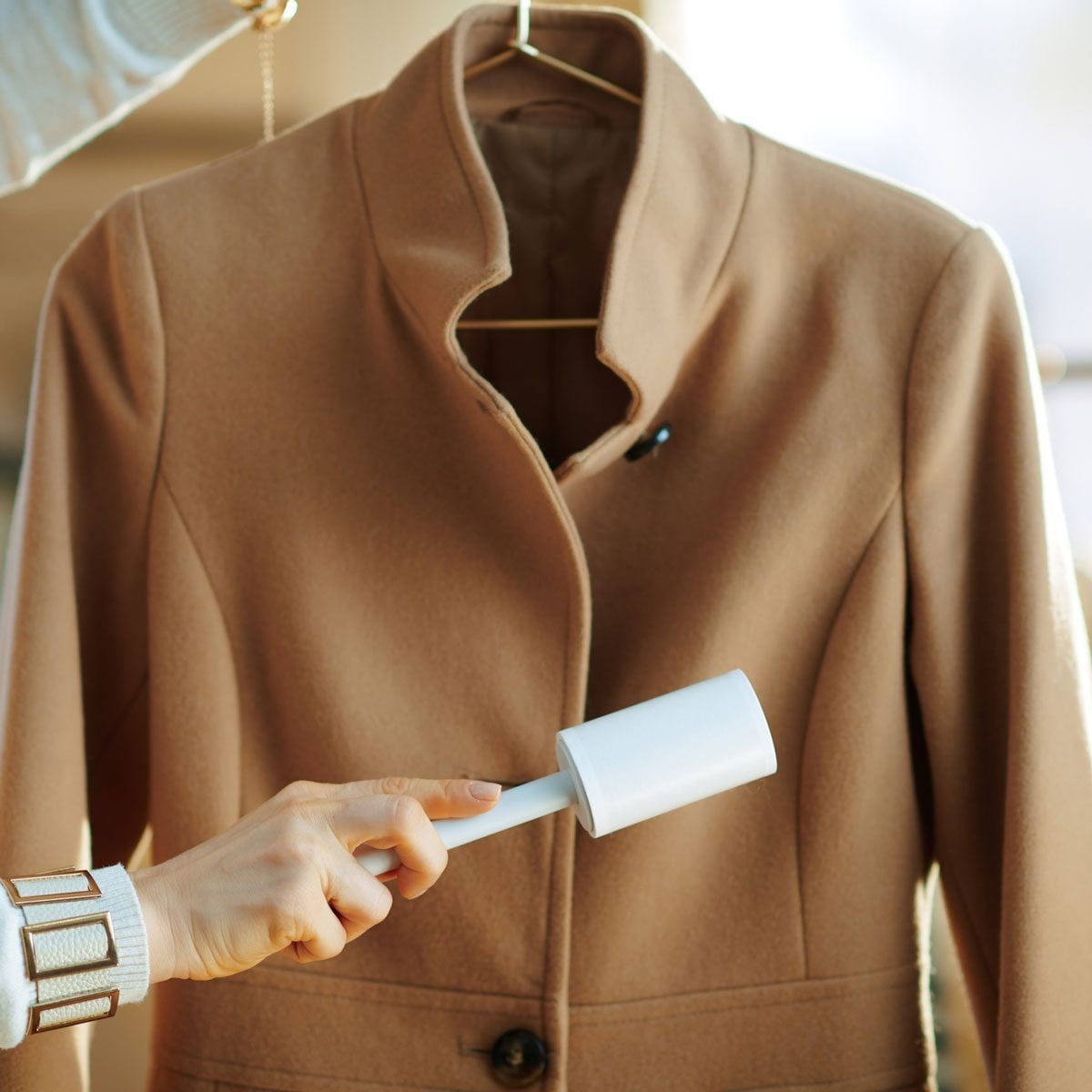 A hand holds a lint roller, removing lint from a brown coat hanging on a wooden hanger in a warm, softly lit indoor environment.