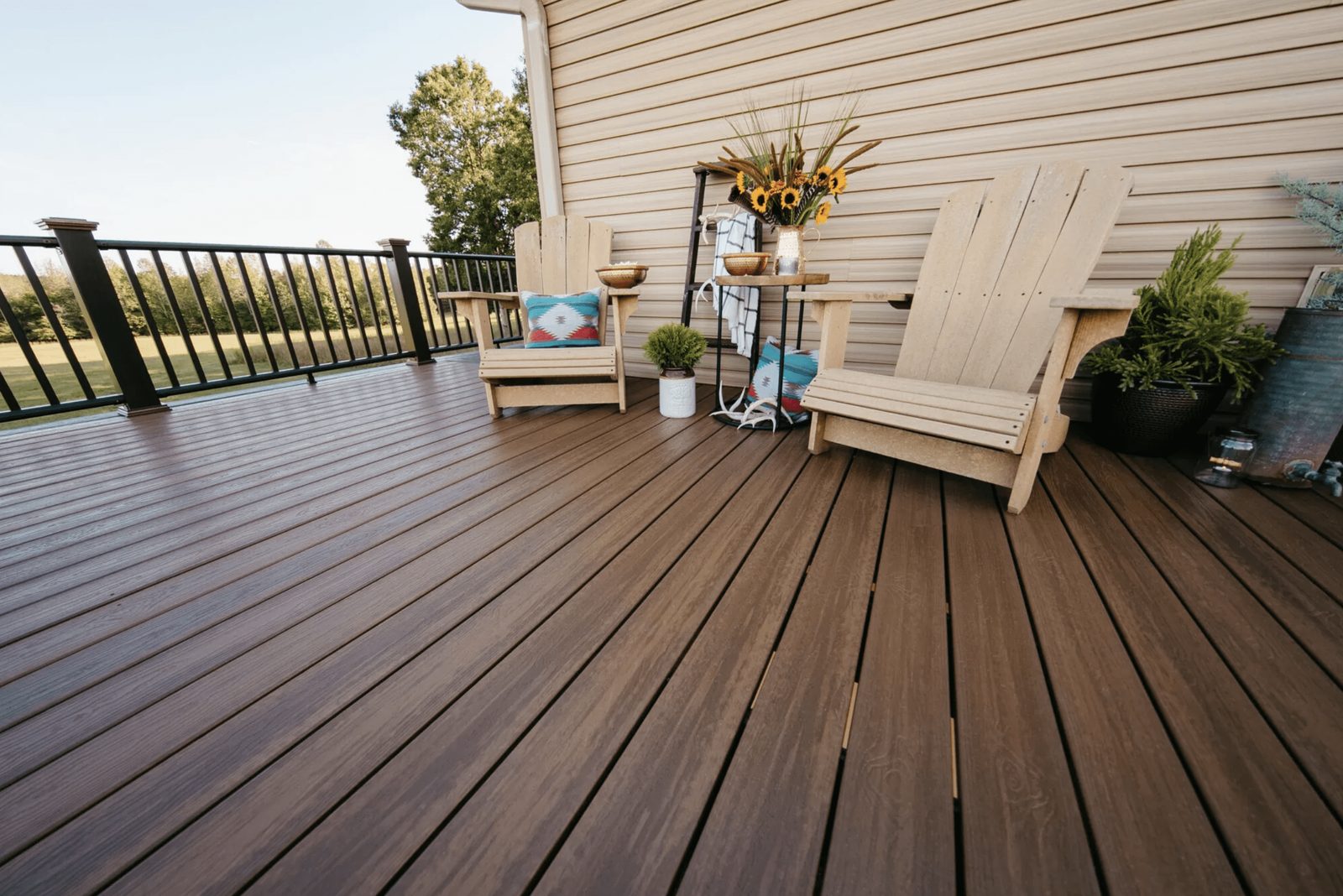 Two wooden adirondack chairs sit on a brown deck, surrounded by potted plants and decorative items, overlooking a green field under a clear
