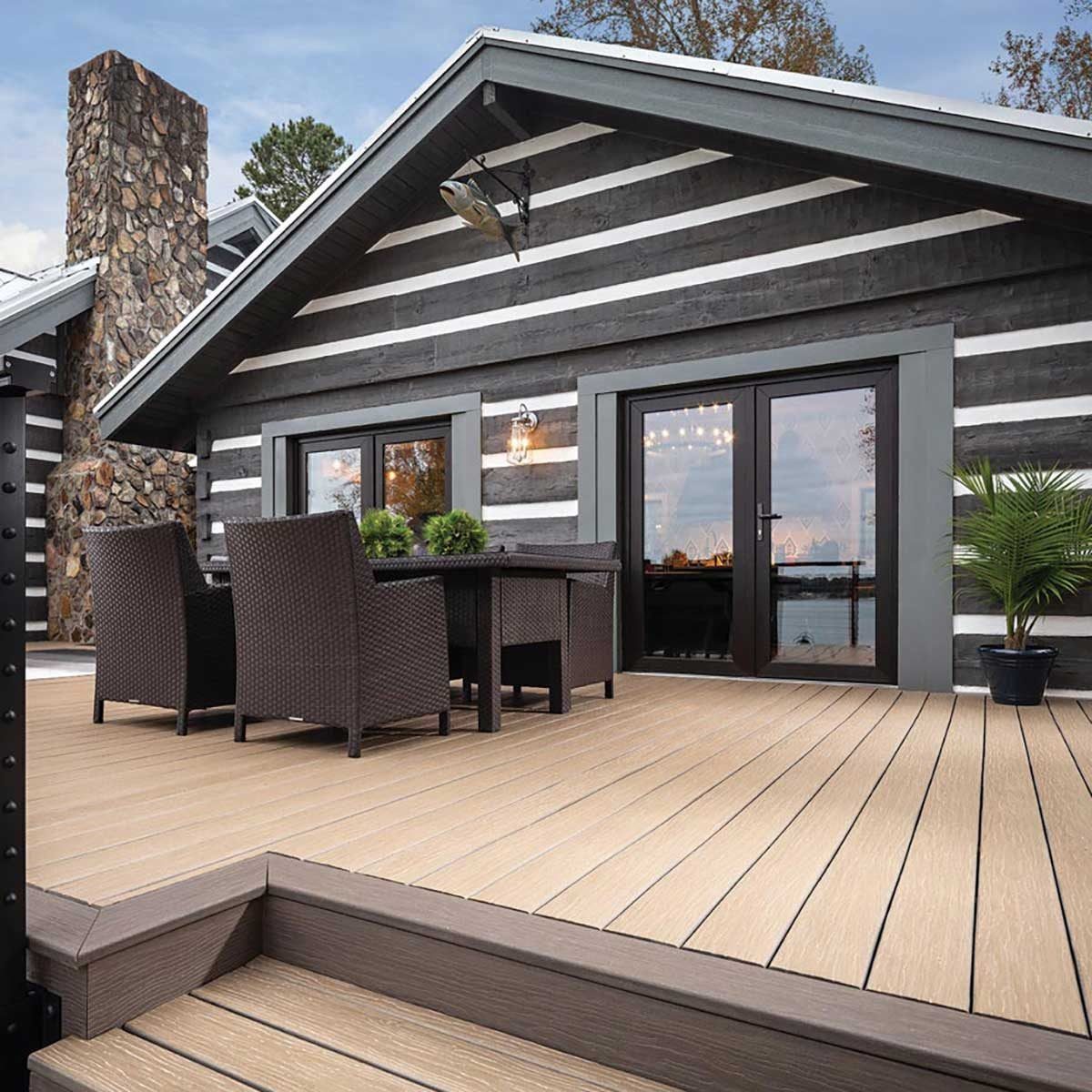 A wooden deck features a table and chairs, set against a log cabin backdrop with large windows, showcasing a peaceful outdoor view by the wa