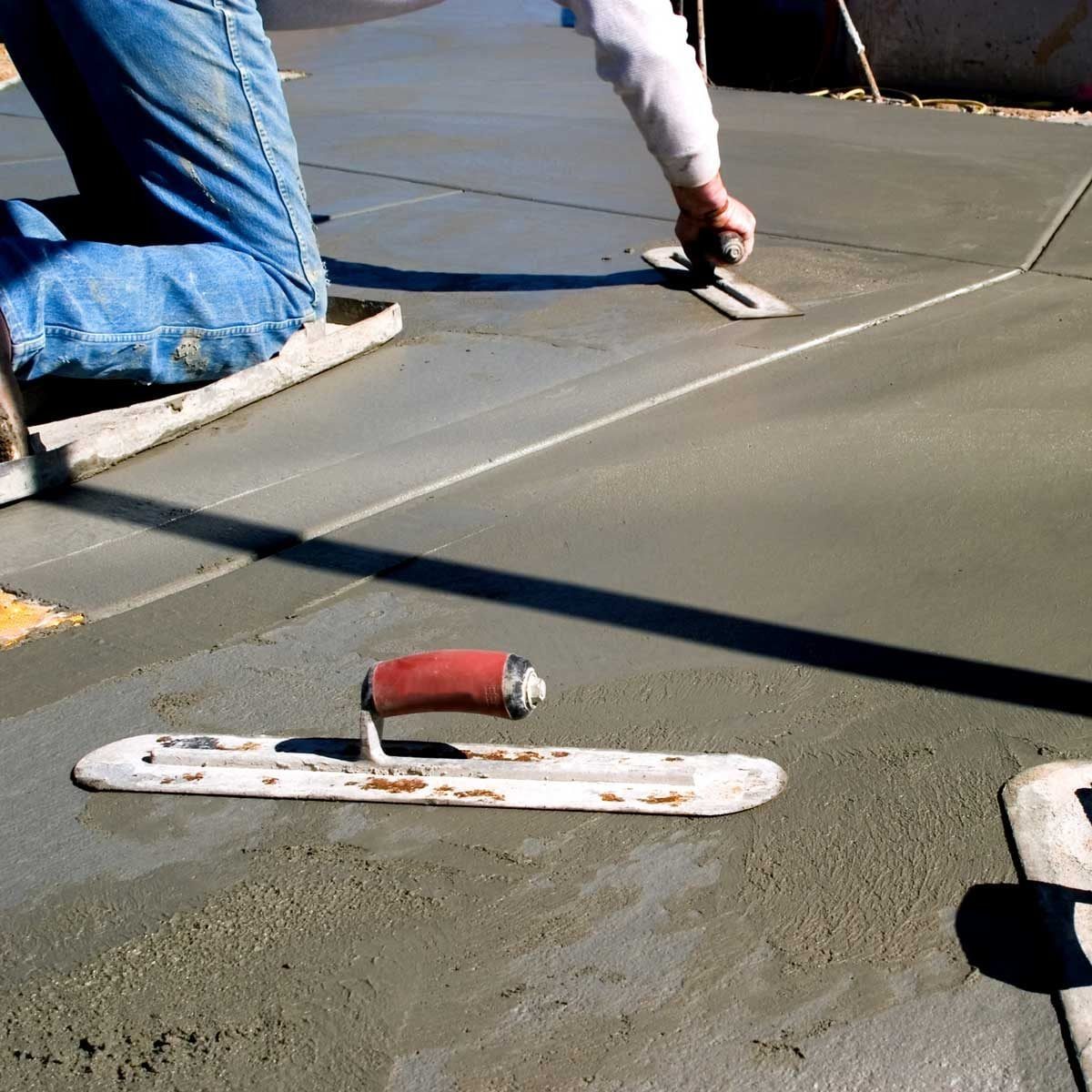 A worker kneels on a fresh concrete surface, smoothing it with a trowel, while another trowel lies nearby on the wet pavement under bright s