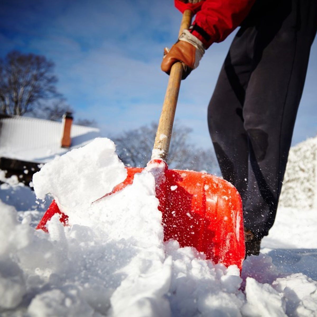 A person uses a red shovel to lift snow, creating a plume of snowflakes in a bright outdoor setting with a clear sky and trees in the backgr