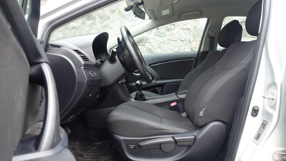 A car interior features a driver's seat and steering wheel facing forward, with the passenger seat beside it, set against a rocky wall outsi