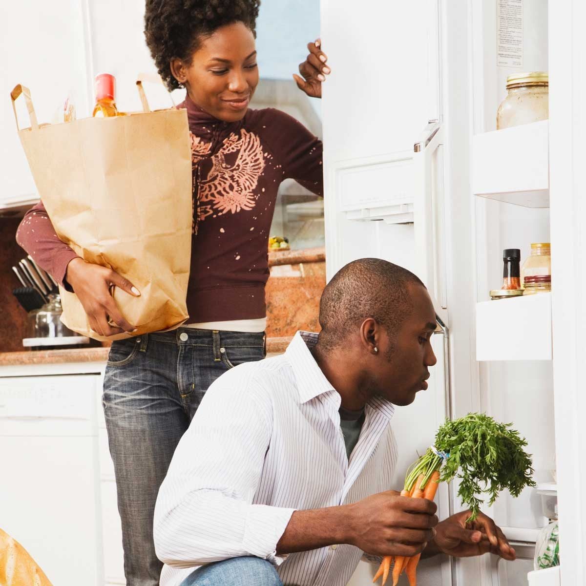 A man kneels by a fridge, holding carrots, while a woman smiles and carries a paper bag full of groceries in a modern kitchen.