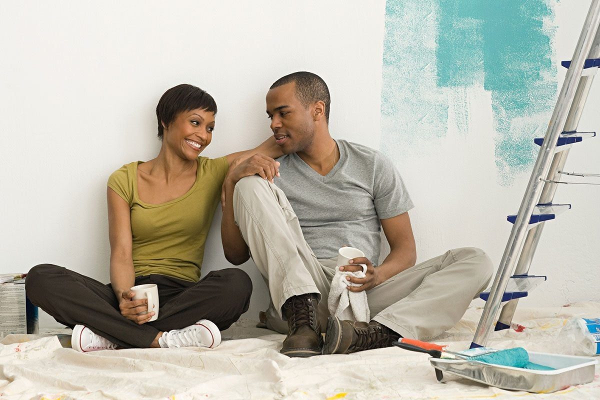 A couple sits on a drop cloth, smiling and sharing a moment, with a partially painted wall and painting supplies nearby.
