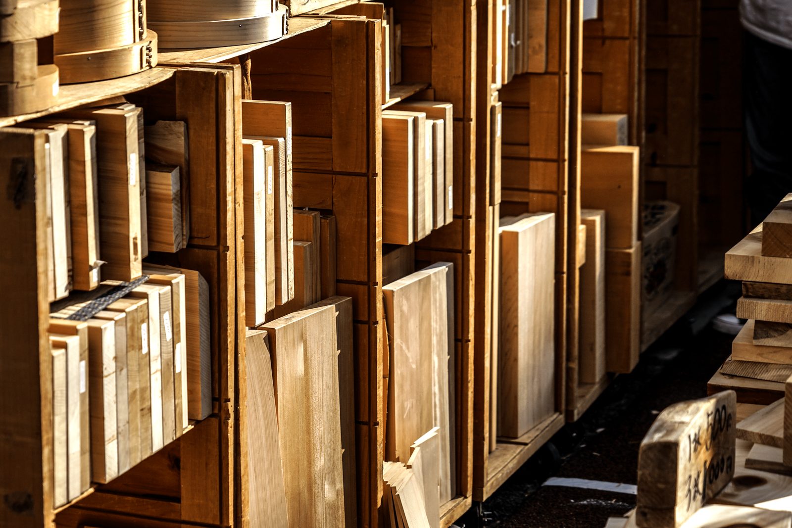Stacks of wooden planks are stored in wooden crates, illuminated by sunlight, in a bustling marketplace setting.