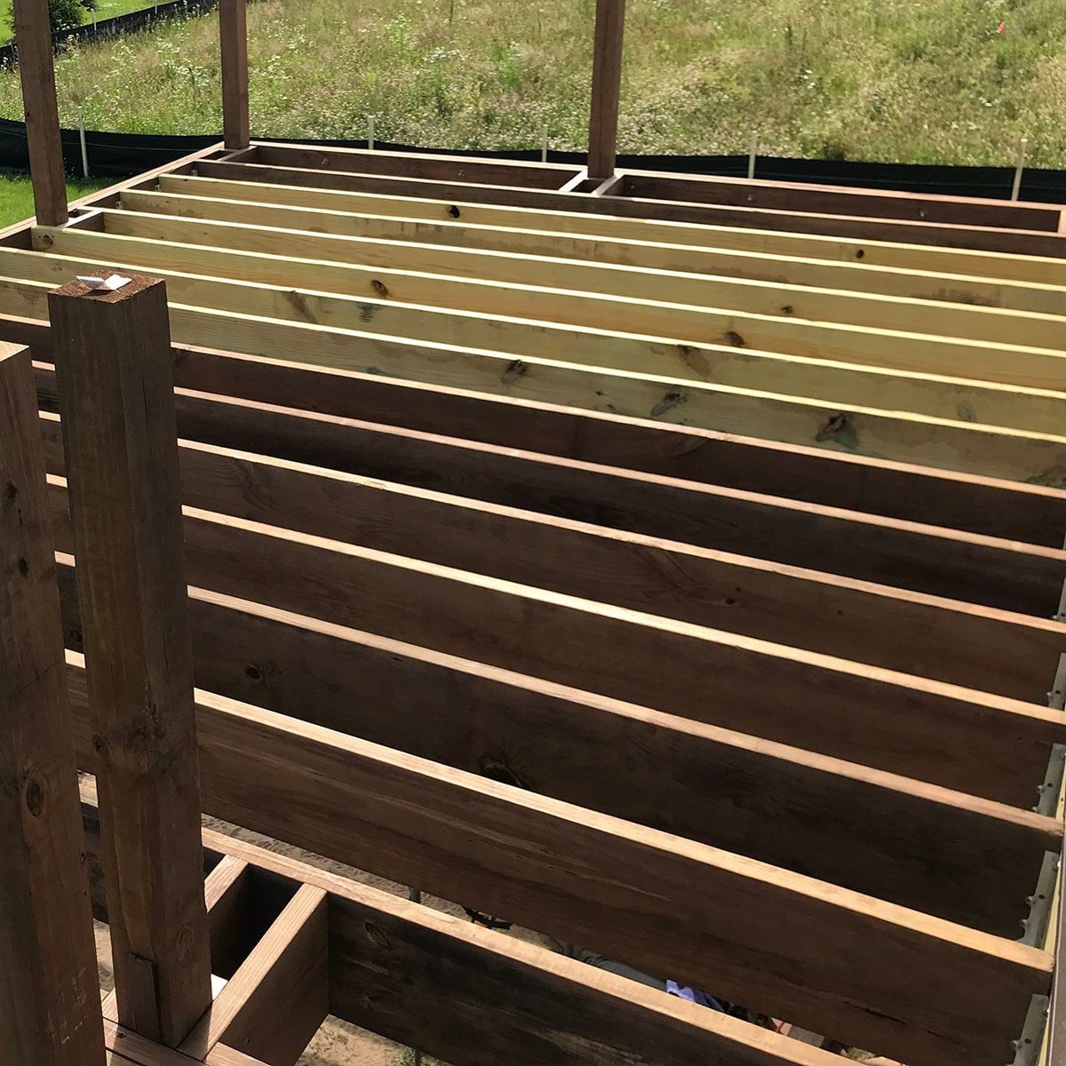 Wooden beams are arranged horizontally, supporting a structure. Green grass and a fence surround the area, indicating an outdoor constructio