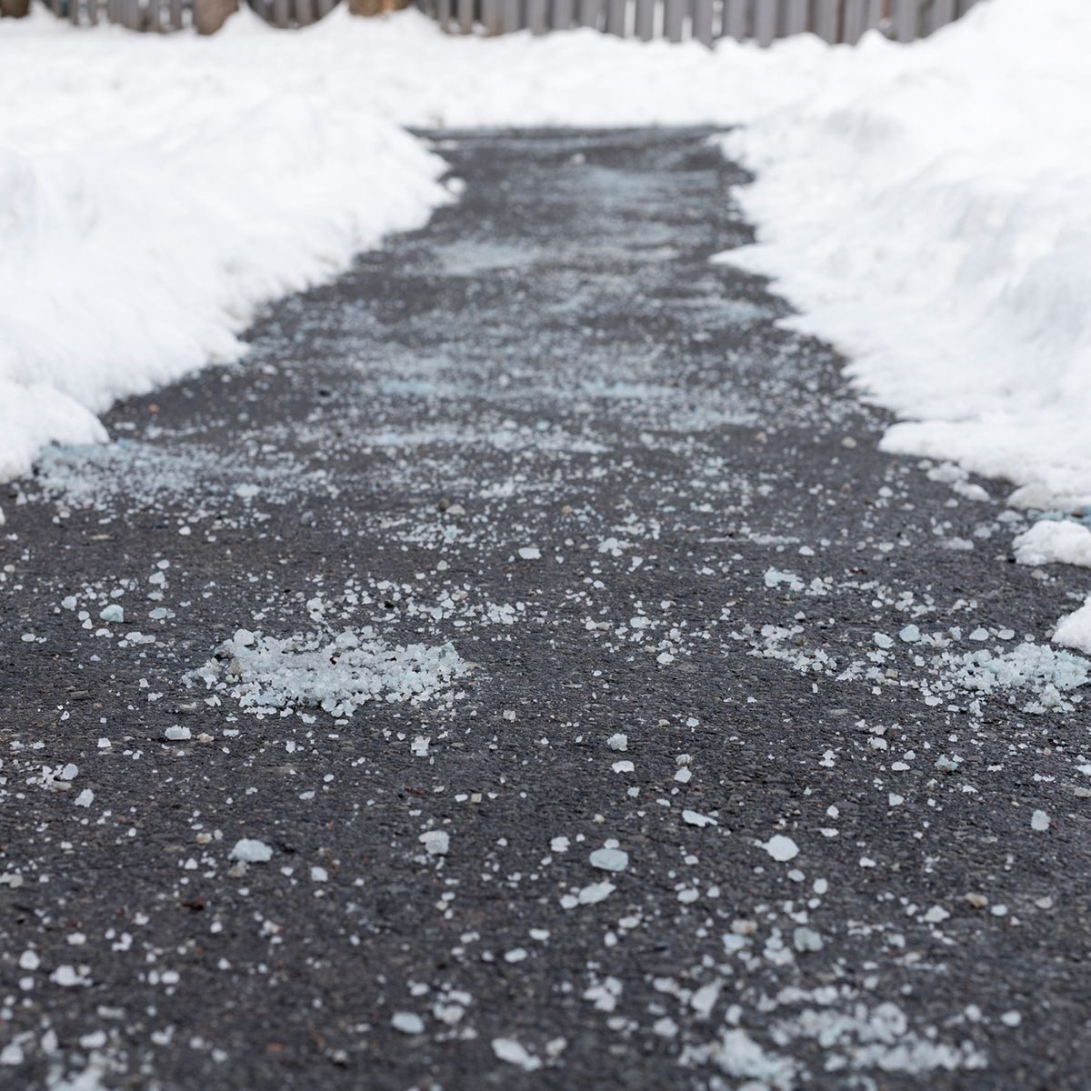 A dark walkway is partially covered in scattered salt, bordered by white snow piles, leading to a wooden fence in the background.