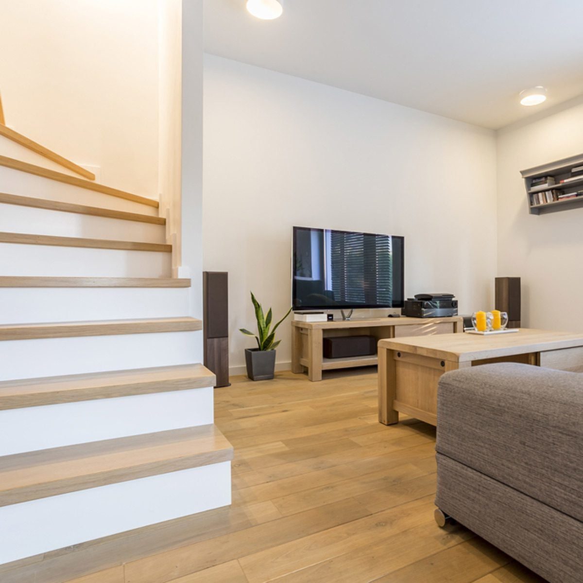A wooden staircase leads to a modern living room featuring a television, coffee table, and potted plant, with warm lighting and wooden floor