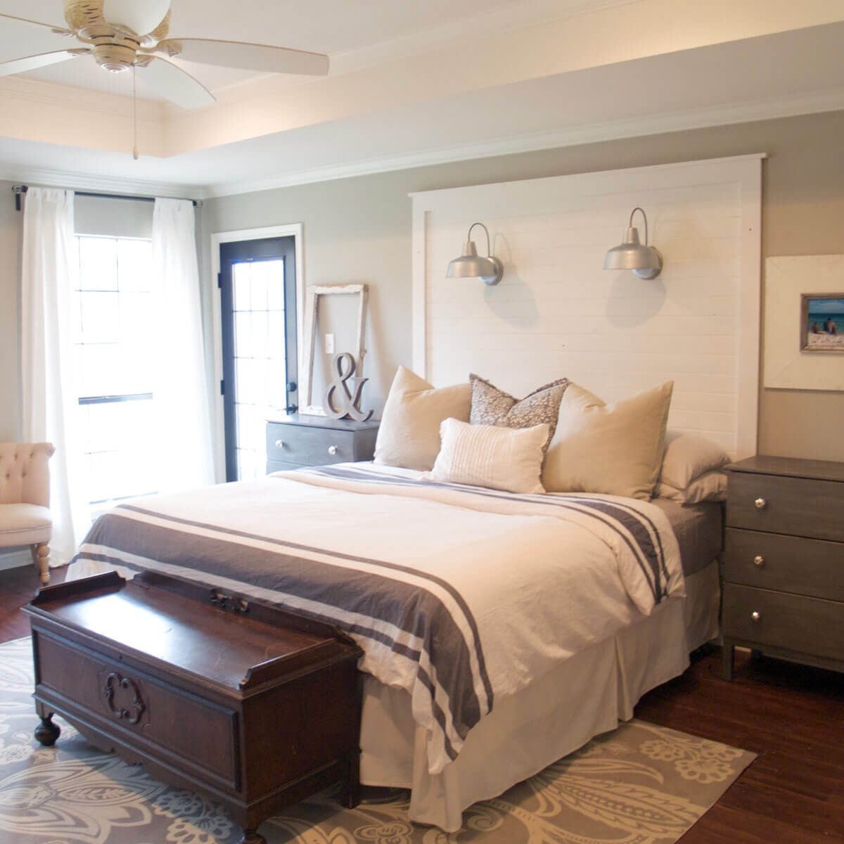 A neatly made bed with decorative pillows rests against a light-colored wall; two nightstands and a vintage trunk are positioned nearby in a