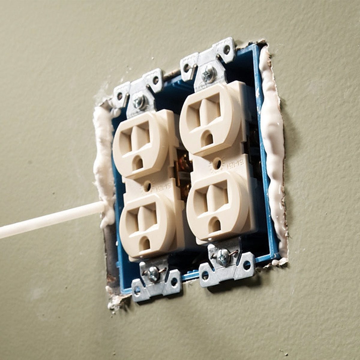 Electrical outlets are installed in a wall, while a tube applies sealant around them to complete the installation. The wall is a muted green