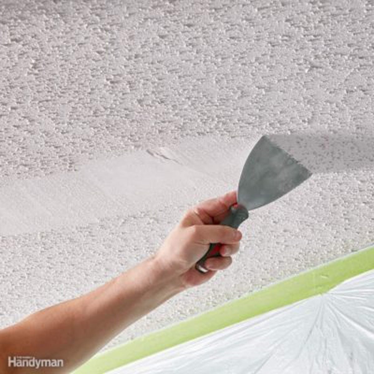 A hand holds a putty knife, smoothing a textured ceiling in a domestic setting, with protective covering along the bottom edges.