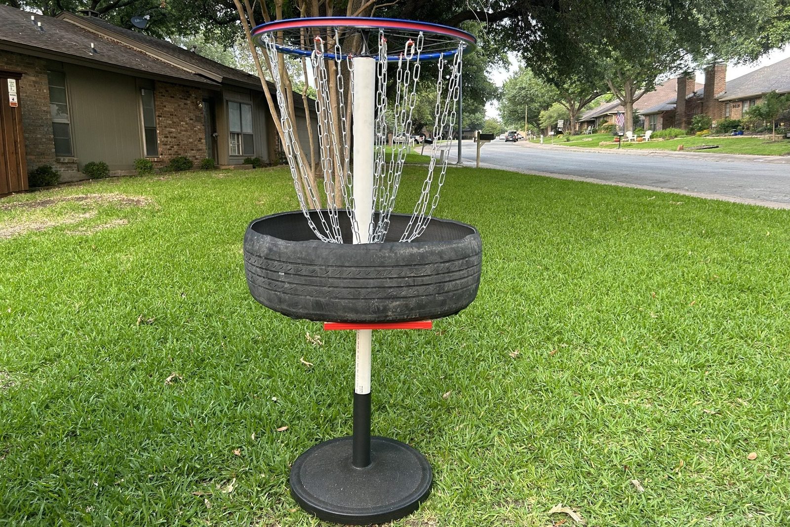 A disc golf basket constructed from a tire and chains stands on a grass lawn, surrounded by residential houses and a quiet street in the bac