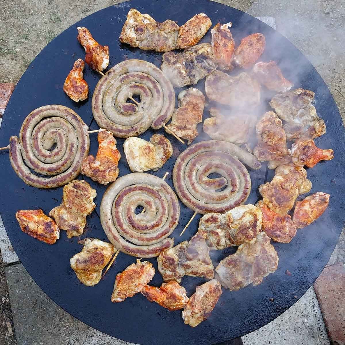 Grilled sausages and chicken pieces sizzle on a round cooking surface, emitting smoke in a grassy outdoor setting.