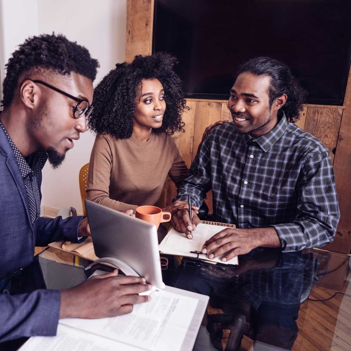 Three people engage in a discussion around a table, reviewing notes and using a tablet, in a bright, modern workspace with wooden accents.