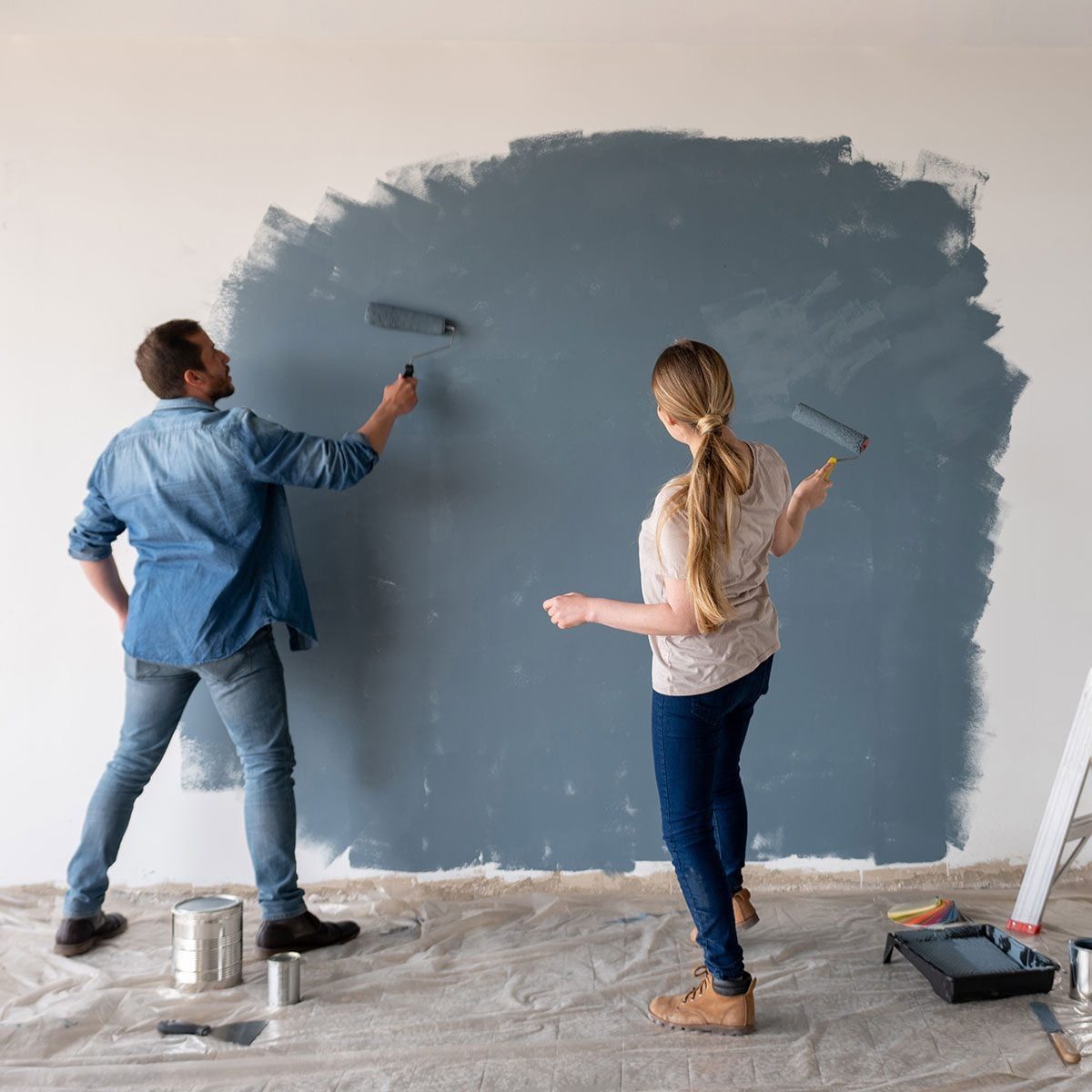Two individuals are painting a wall with a roller. A tarp covers the floor, and paint supplies are nearby in a well-lit indoor setting.