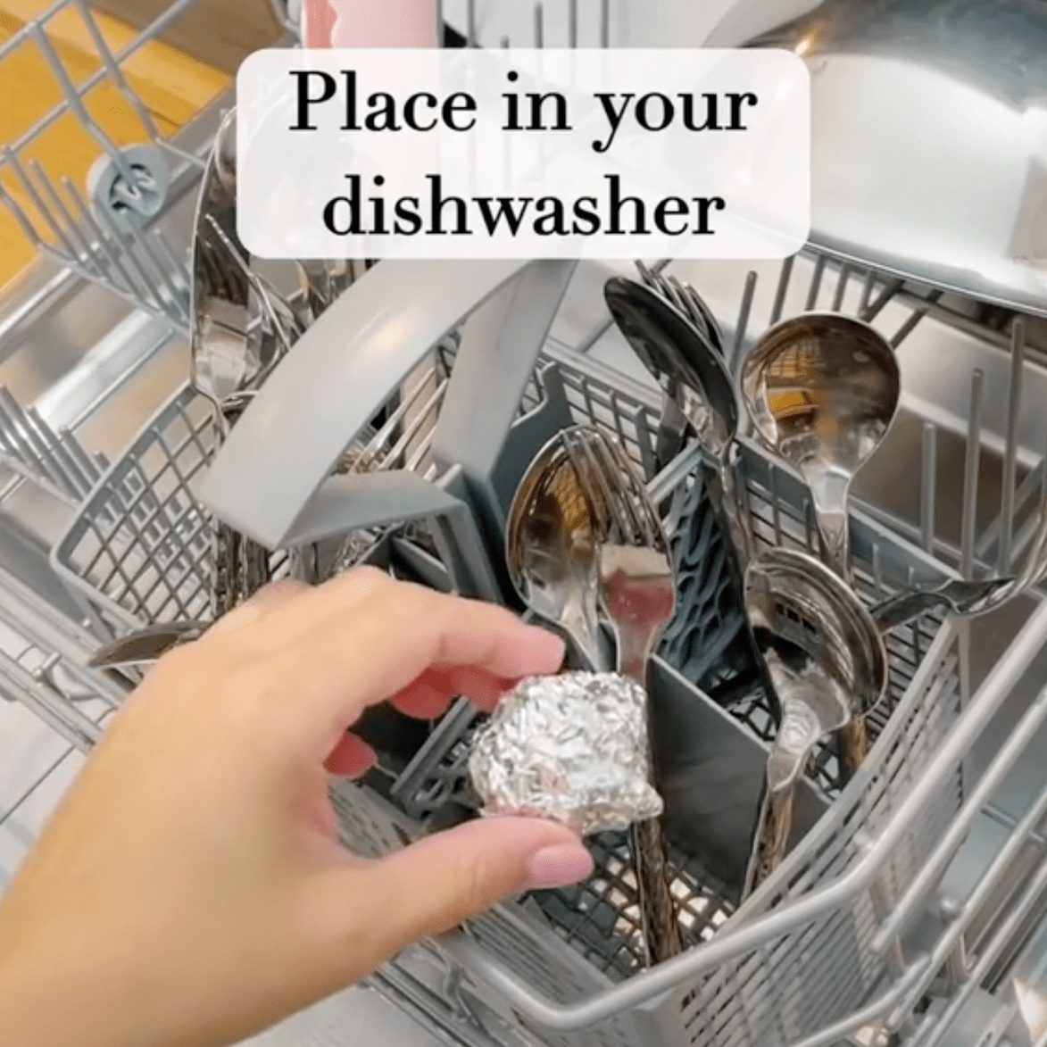 A hand holds a crumpled piece of foil, placing it into a dishwasher filled with various utensils, including spoons and forks.