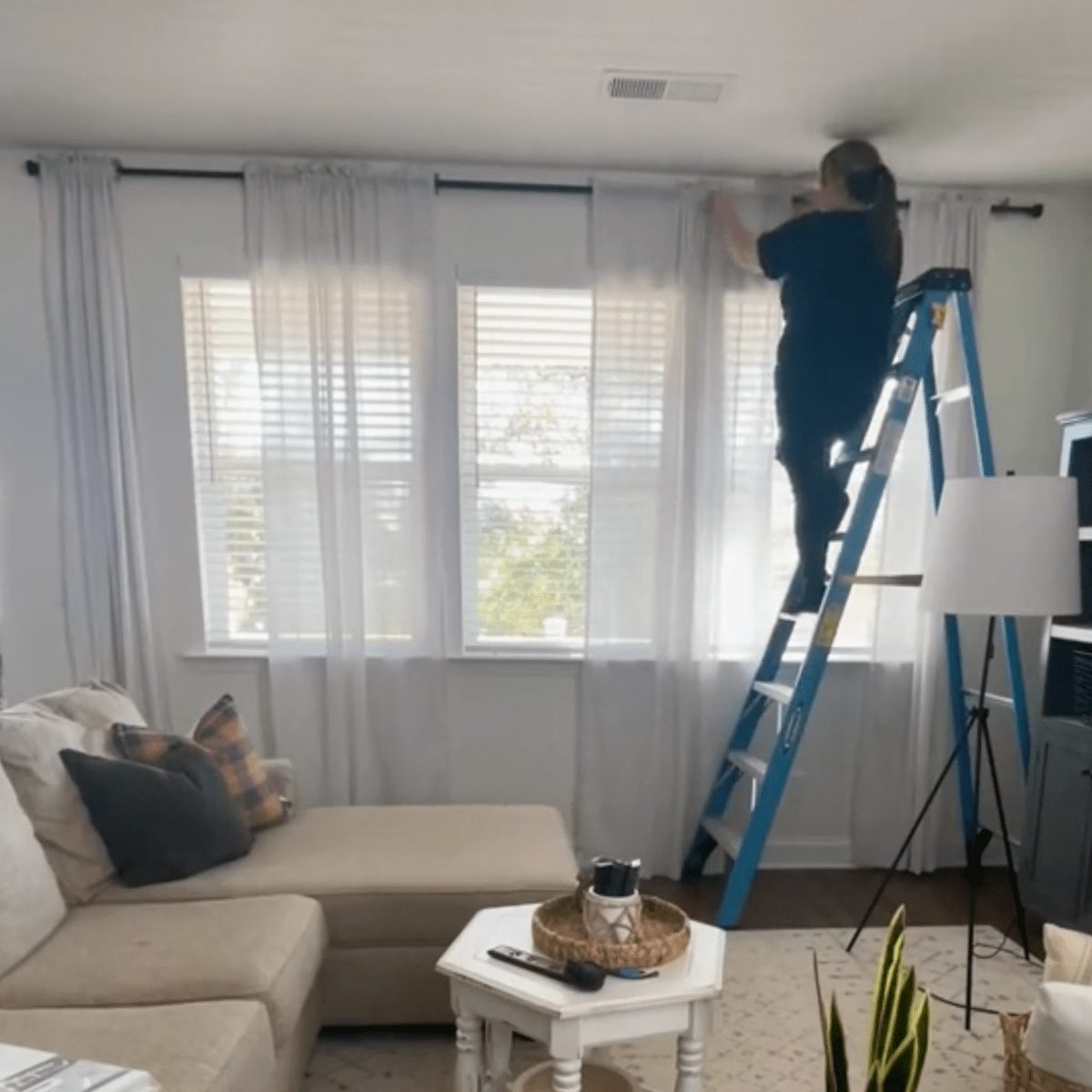 A person is adjusting curtains while standing on a blue ladder, in a bright living room with large windows and light-colored furniture.